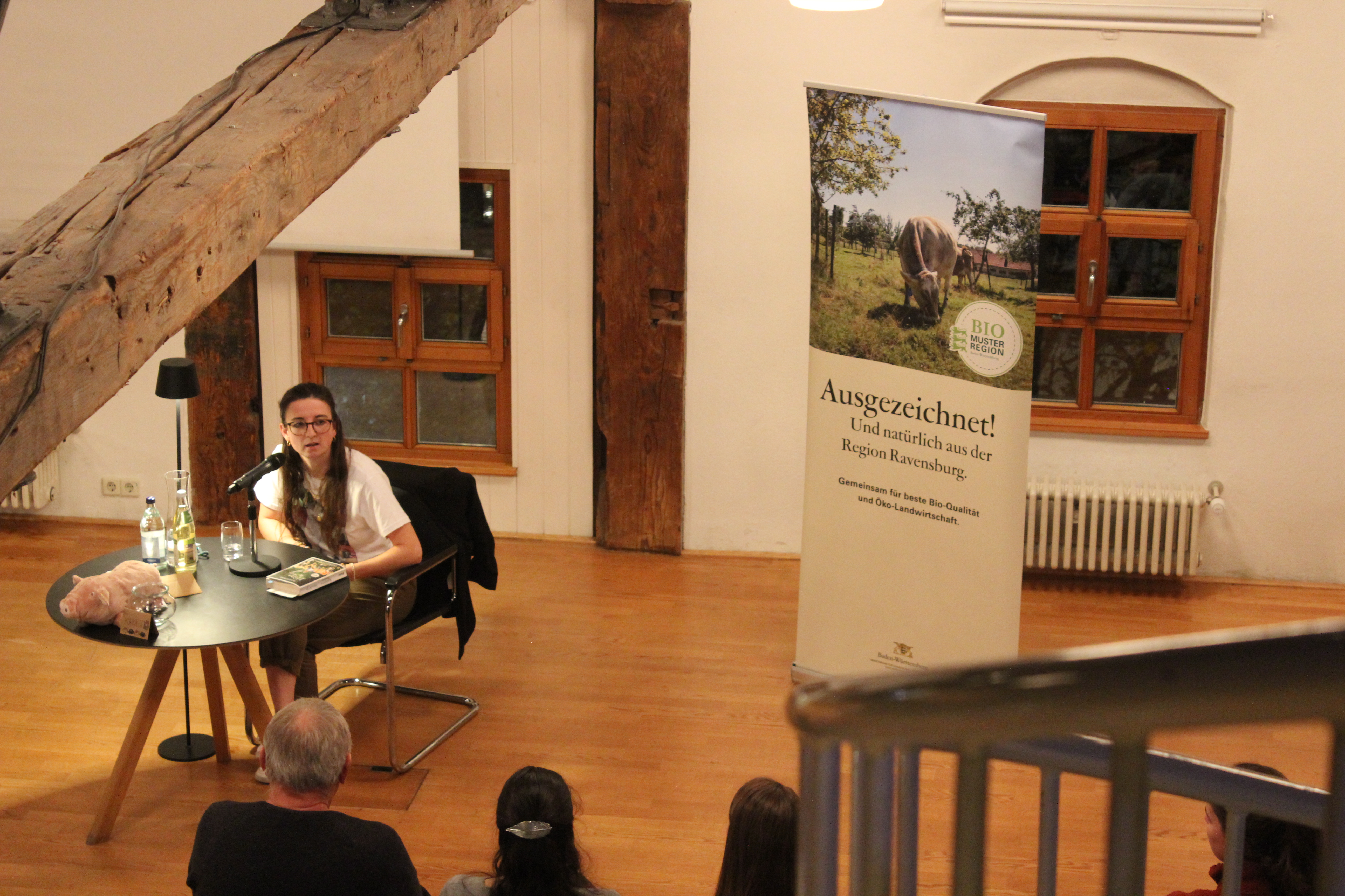 Madeleine Becker am Tisch mit ihrem Buch, sie spricht mit dem Publikum. im Hintergrund das Rollup der Bio-Musterregion