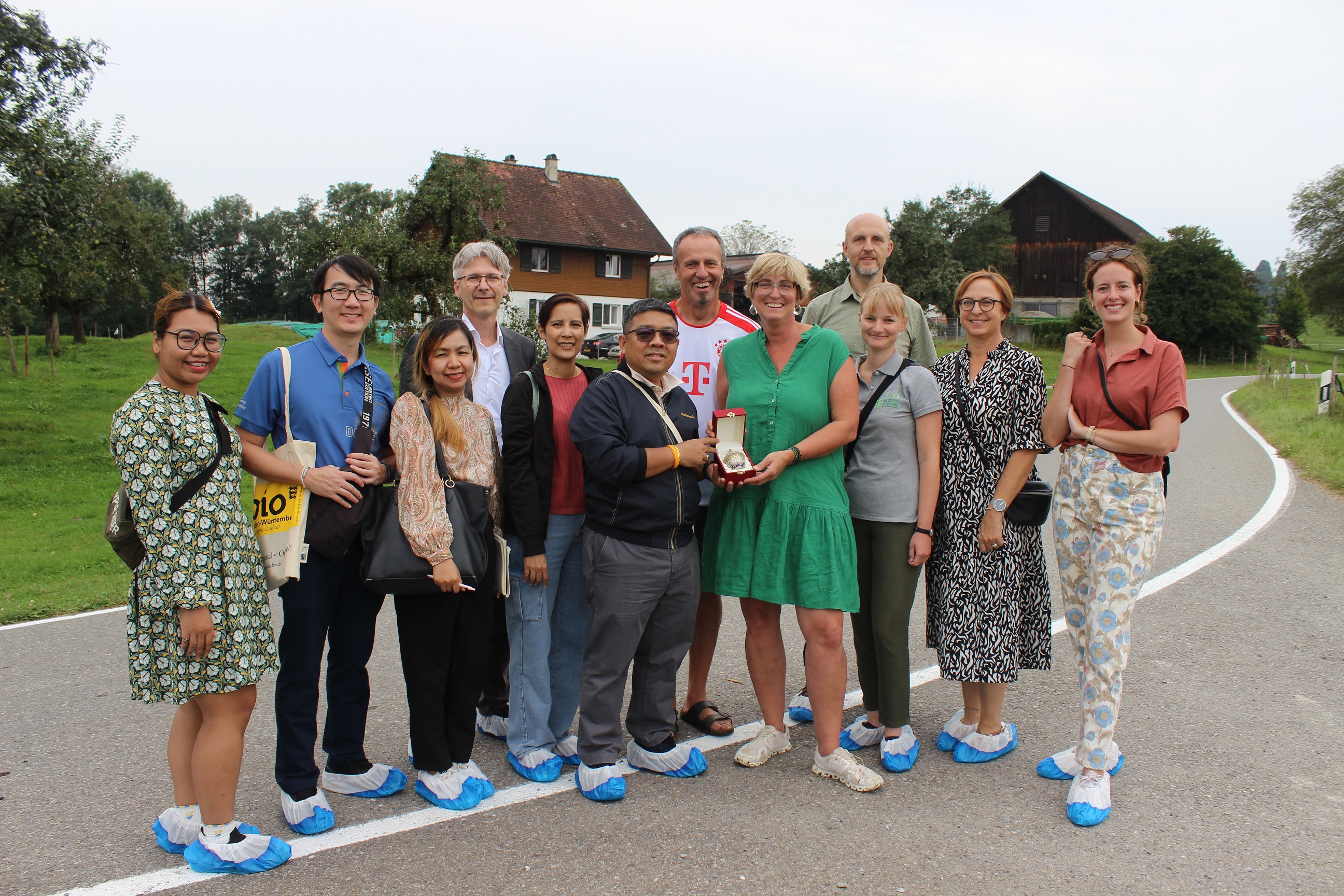 Zu sehen sind die Personen der Delegation aus Thailand beim Biohof Fäßler mit der Streuobstwiese und eine Wohnhaus im Hintergrund. Mit dabei ist auch ein Teil der Lenkungsgruppe