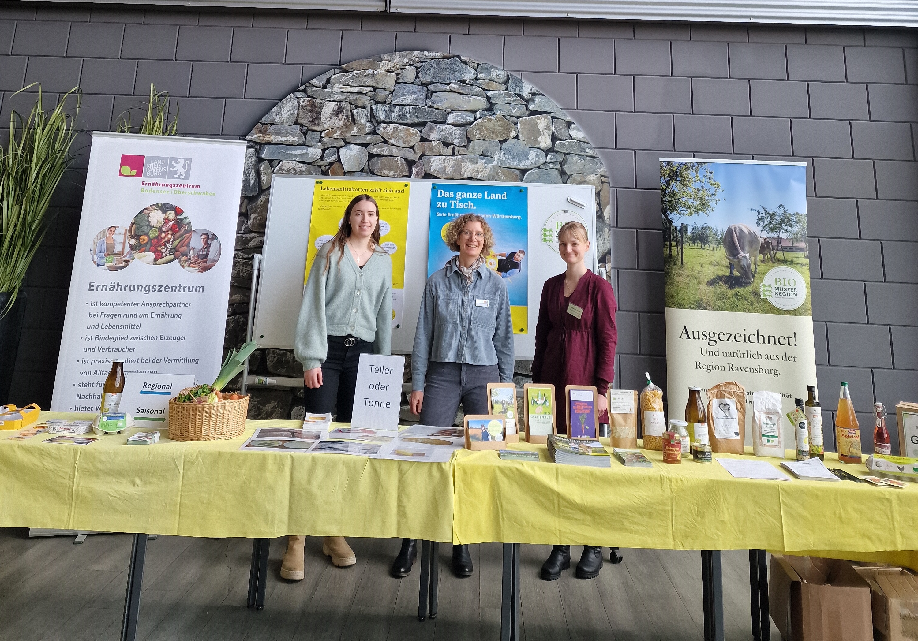Drei Frauen stehen bei ihrem Infostand. Zu sehen sind Roll-ups mit den Logos des Ernährungszentrums und der Bio-Musterregione. Auf den Tischen vor ihnen stehen Produkte und Infomaterial. 