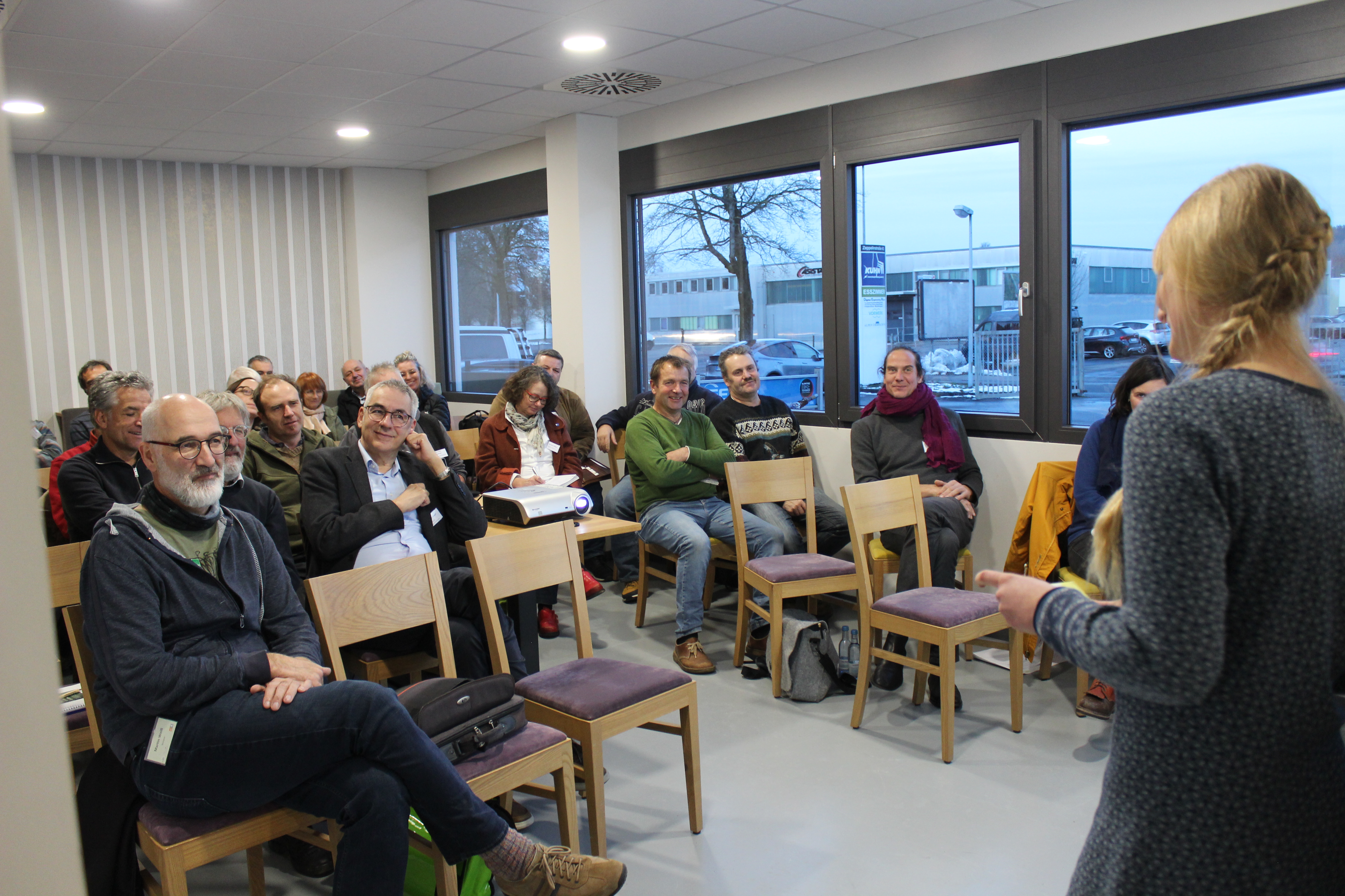 Katharina Eckel steht im Vordergrund den Rücken zum Betrachter und spricht vor den Teilnehmeri/innen des Bio-Forums. Diese lauschen zum Großteil mit einem Lächeln im Gesicht. 