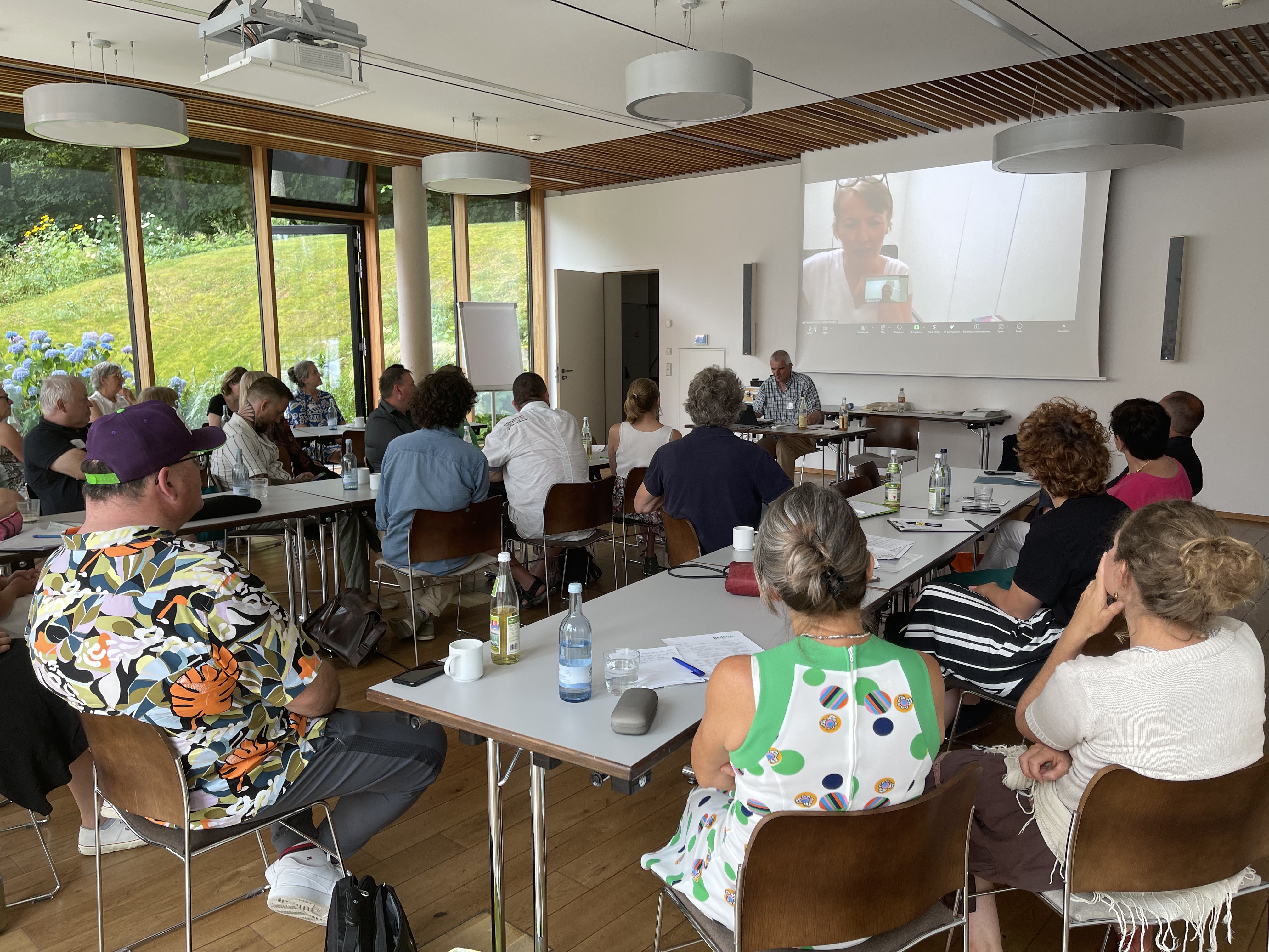 Teilnehmerinnen und Teilnehmer sind sitzend von hinten zu sehen. Alle schauen auf die Leinwand  auf der das Bild von Verena Fiedler zu sehen ist, die spricht