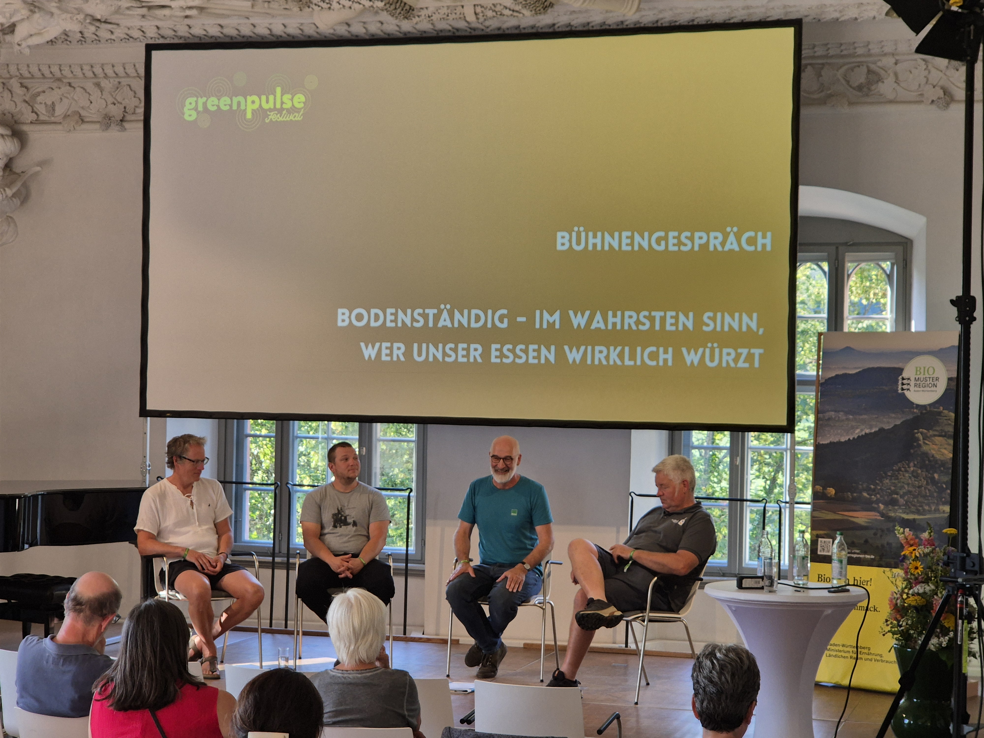 Man sieht die Bühne, im Hintergrund steht auf der Leinwand der Titel des Bühnengesprächs, davor sitzen vier Männer auf Stühlen