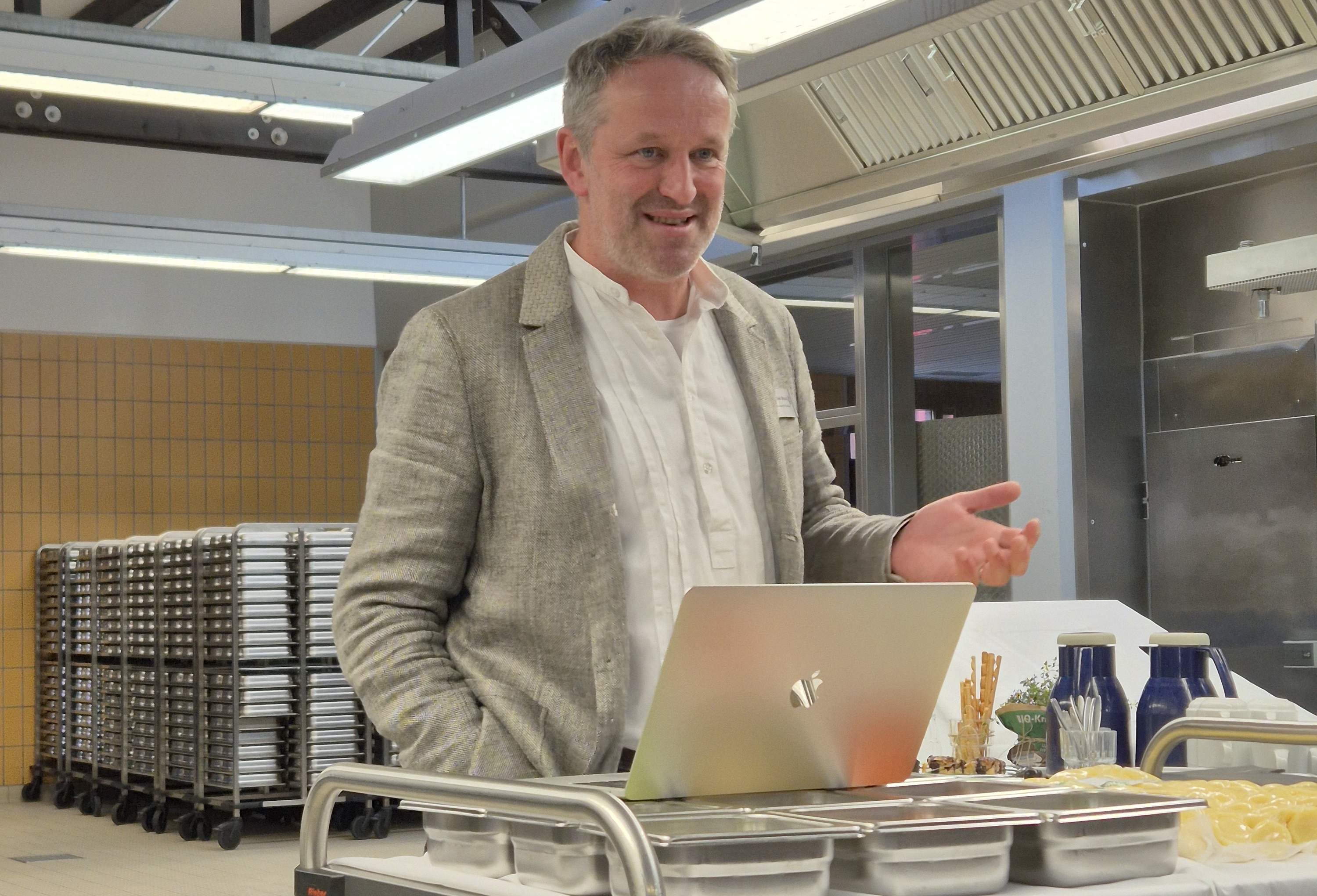 Ein Mann (Karsten Bessai) steht vor einem Laptop, schaut ins Publikum und referiert. In Hintergrund sieht man die Großküche.