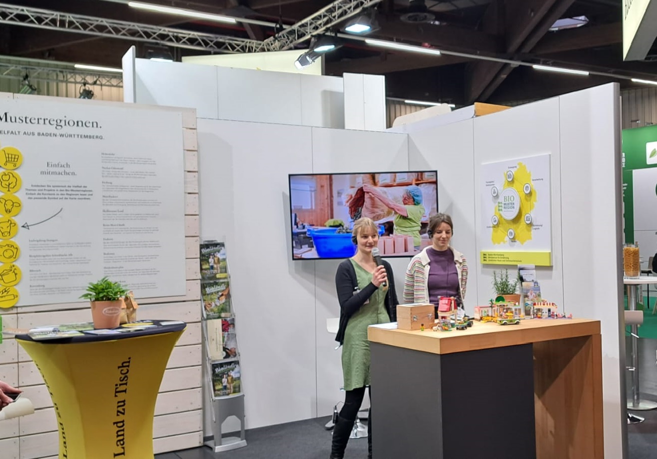 Zwei junge Frauen an der Theke des Gemeinschaftsstandes der Bio-Musterregionen. Eine davon spricht in ein Mikrofon.