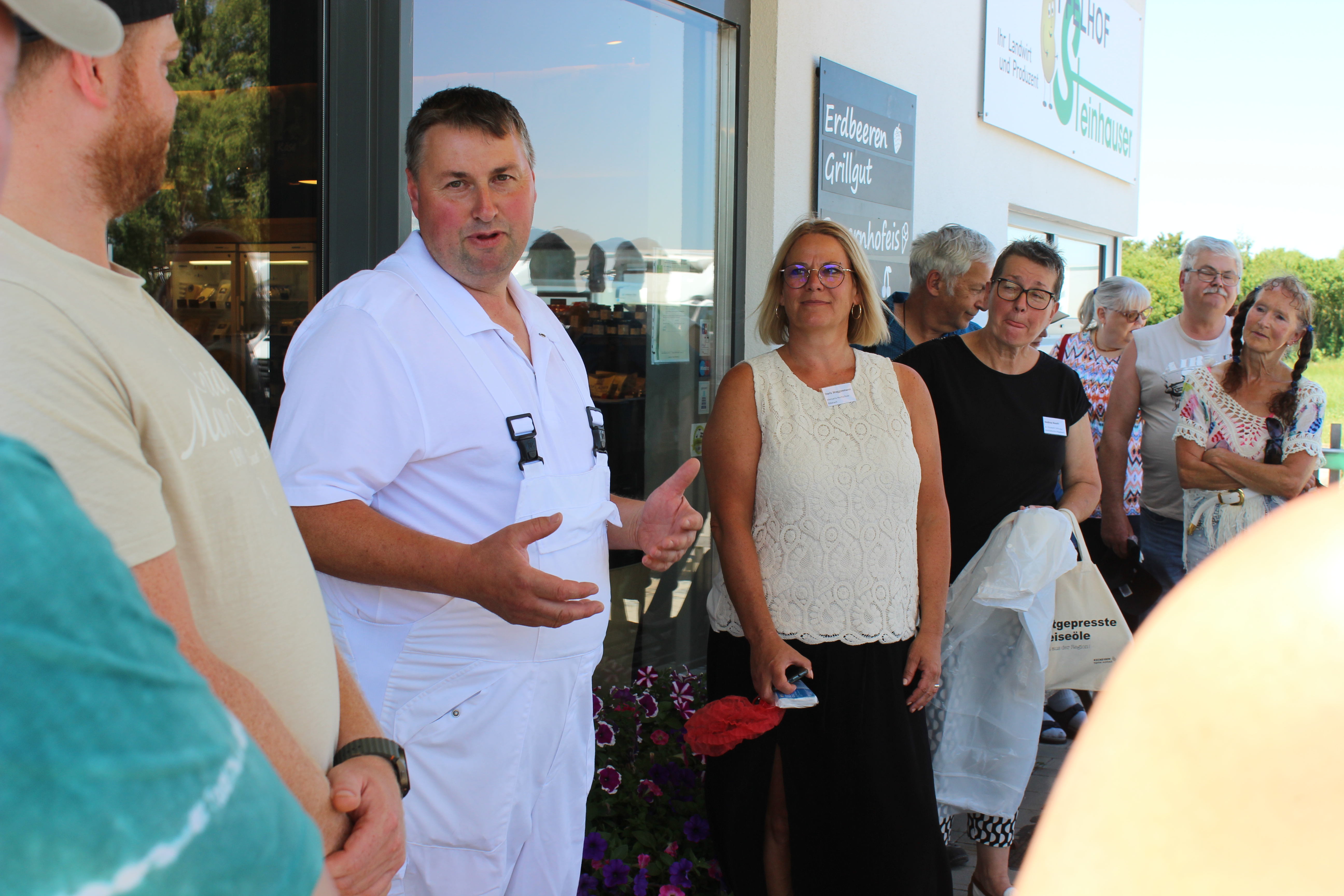 Ein Mann (Franz Steinhauser) steht in weißer Arbeitskleidung mittig und erzählt. Im Hintergrund sieht man Teilnehmerinnen und Teilnehmer die aufmerksam zuhören