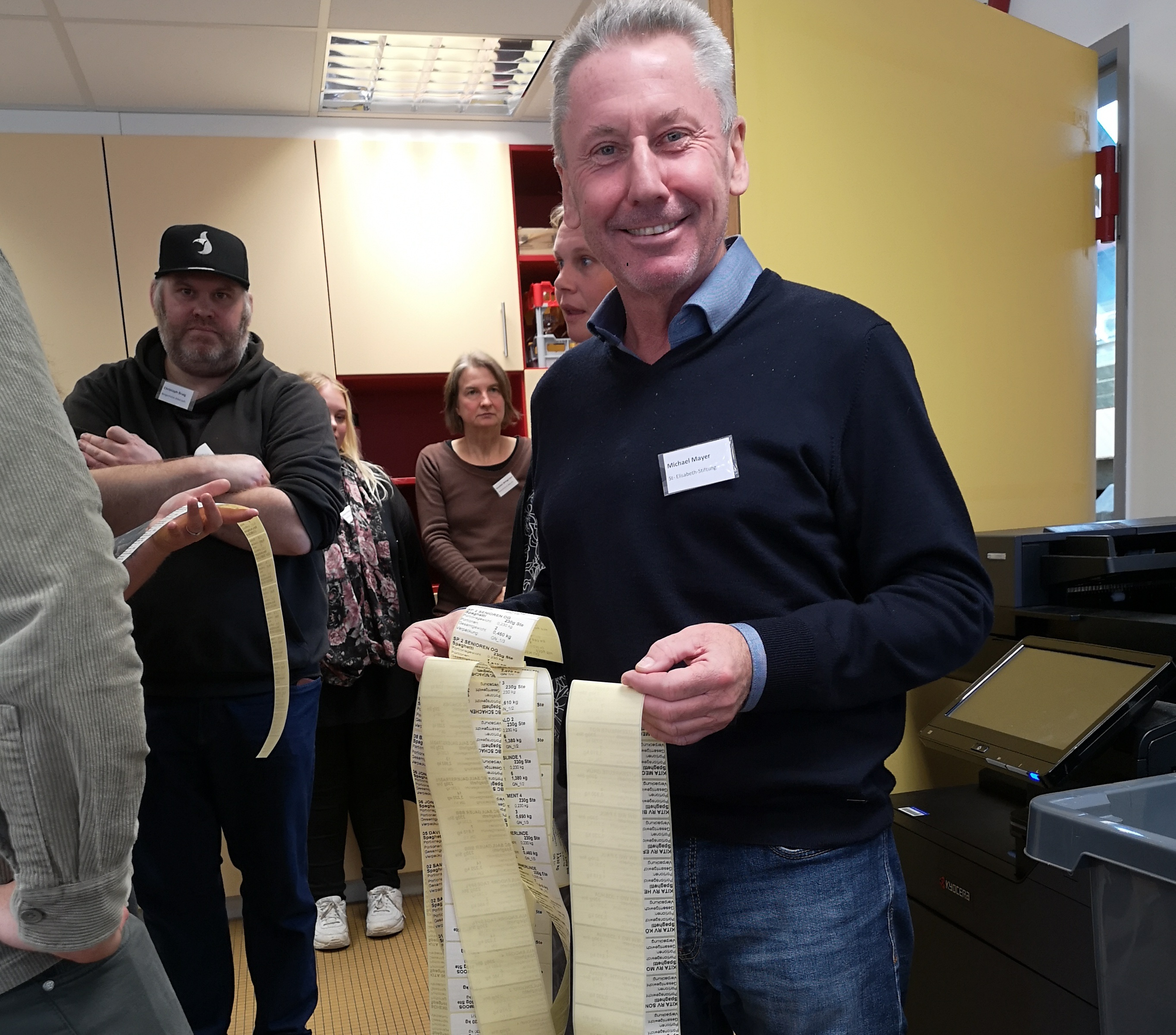 Der Betriebsleiter lächelt in die Kamera und hält lange Bahnen mit bedruckten Etiketten in den Händen. Im Hintergrund sieht man einige Teilnehmende.