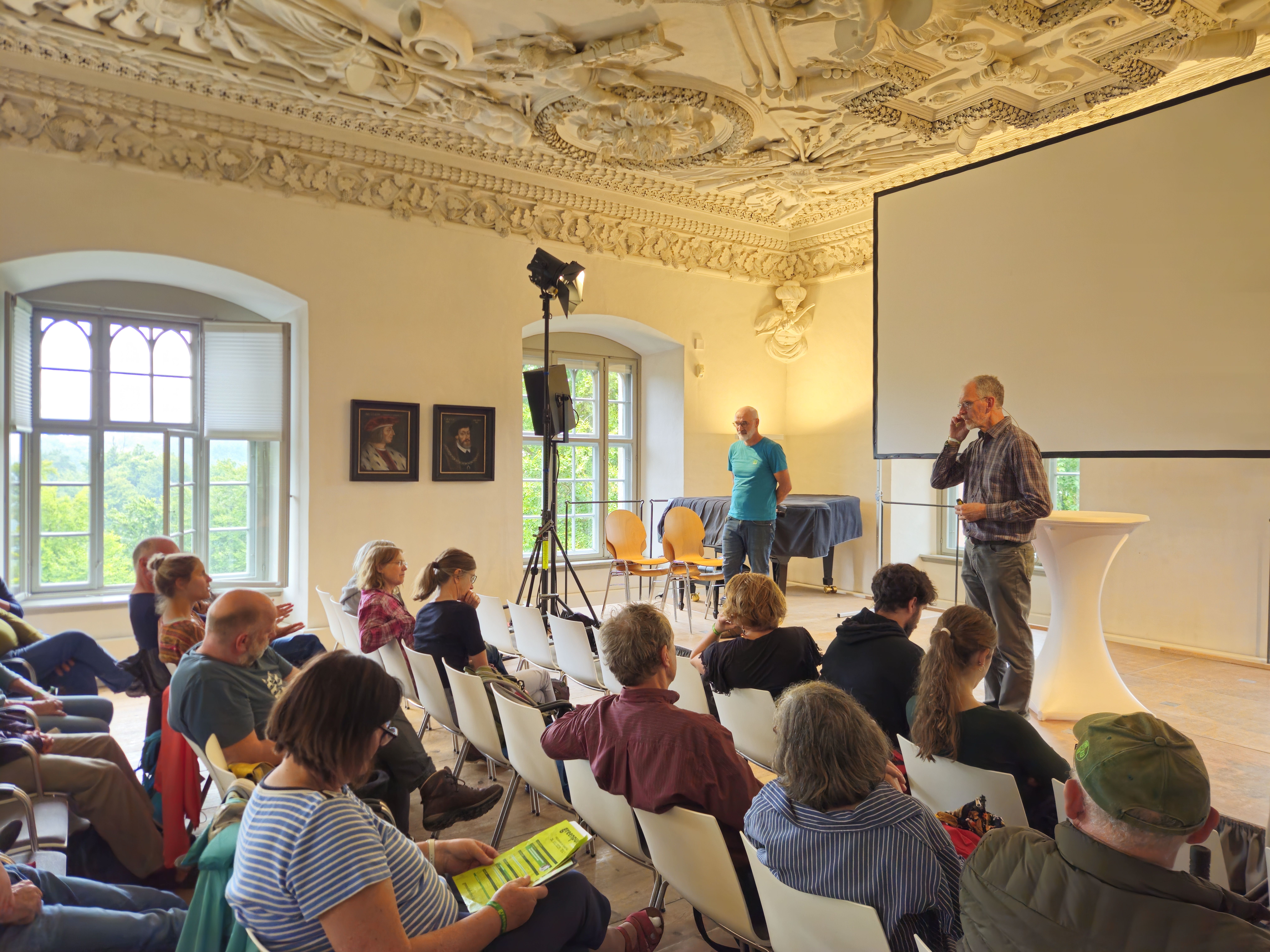 Zwei Männer stehen auf der Bühne in einem Saal und davor sieht man von hinten die Zuhörer