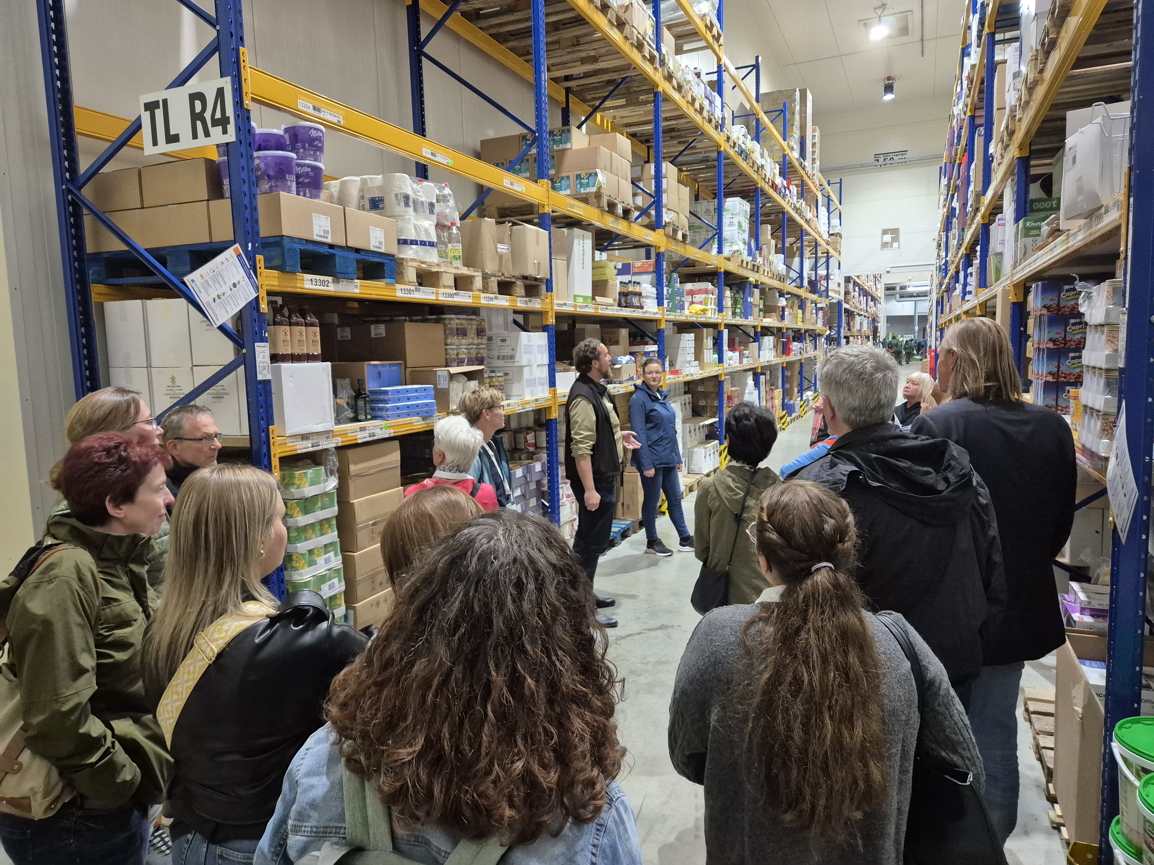 Teilnehmende stehen in einem Gang zwischen den Regalen im Lager von Früchte Jork. Maximilian Jork erklärt sein System. 
