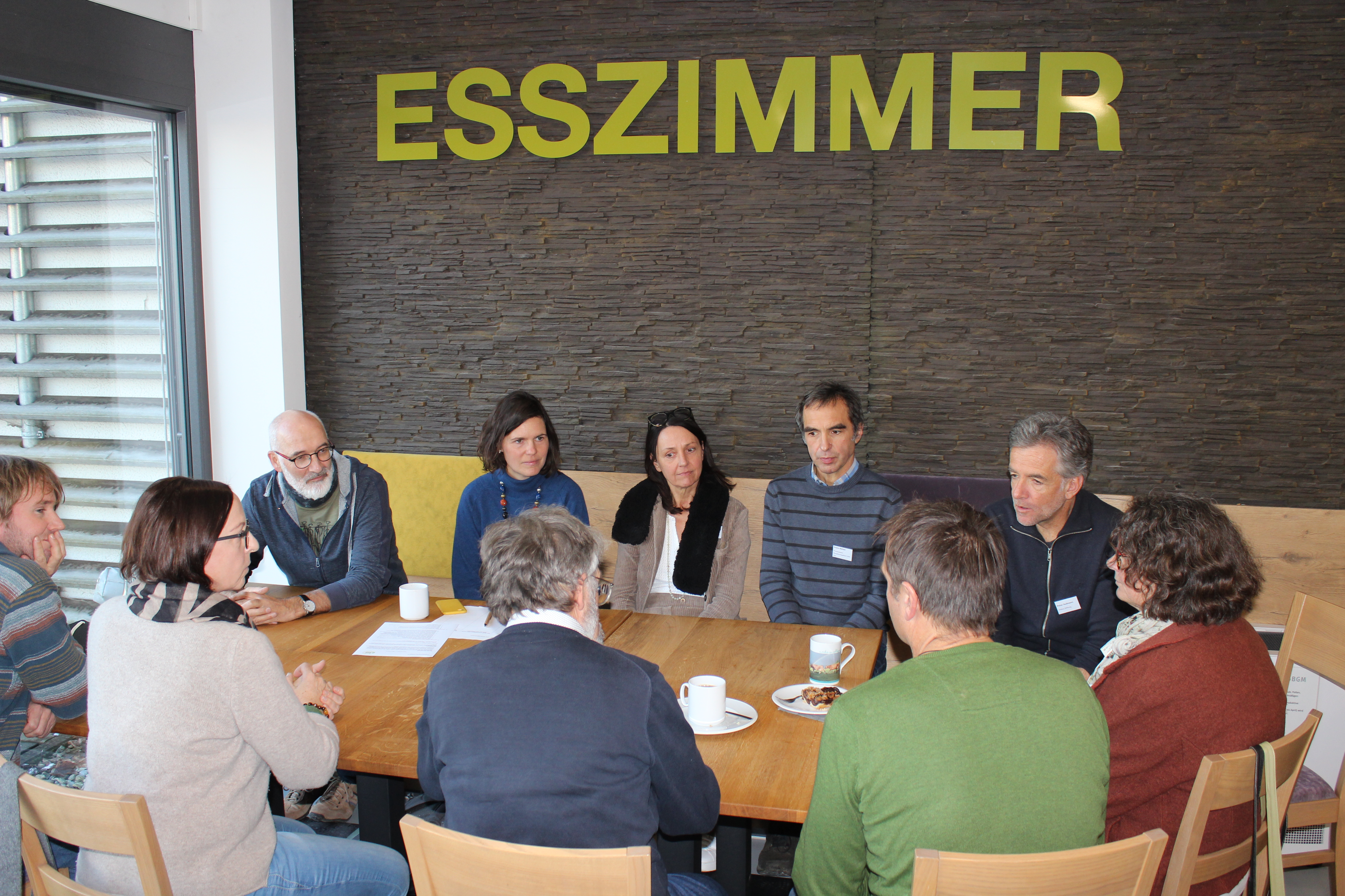 Man sieht einige Teilnehmer/innen um einen Tisch beim Workshop. Im Hintergrund prangen die Lettern 