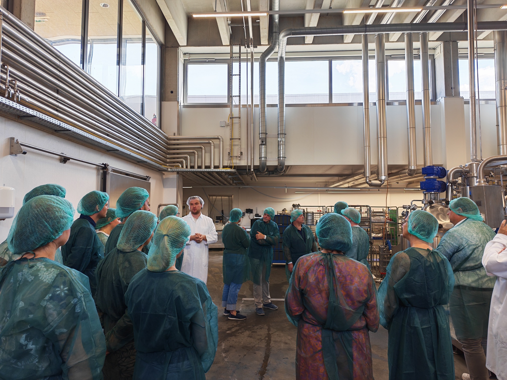 Man sieht die Gruppe mit Hygieneüberkleidung im Kreis stehen. Ein Mann im weißen Kittel spricht. Im Hintergrund sieht man die Produktionsanlagen