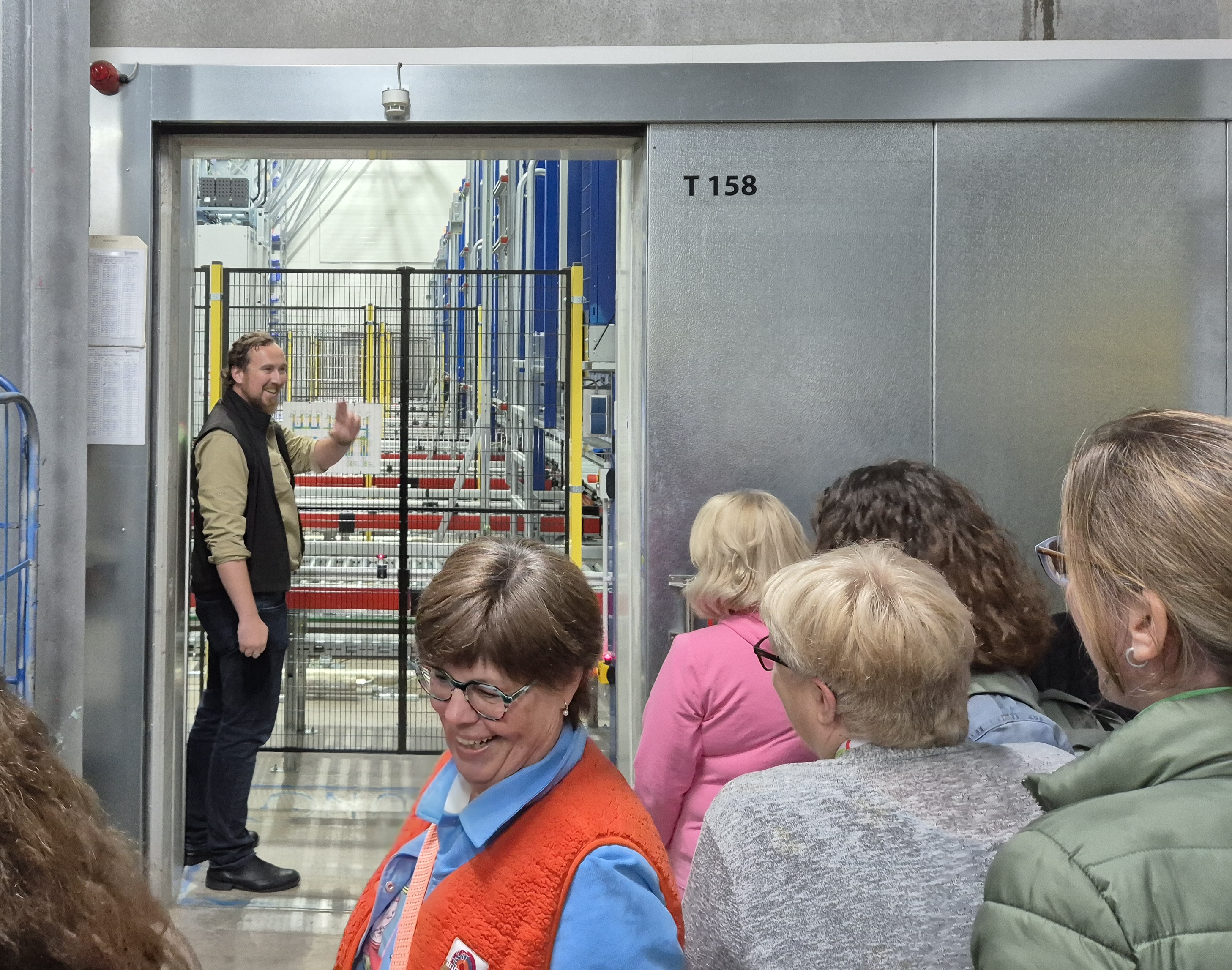 Maximilian Jork steht mit dem Gesicht zu den Teilnehmenden am vergitterten Tor hinter dem man verschiedene Transportbänder aus dem Tifkühllager sieht. 