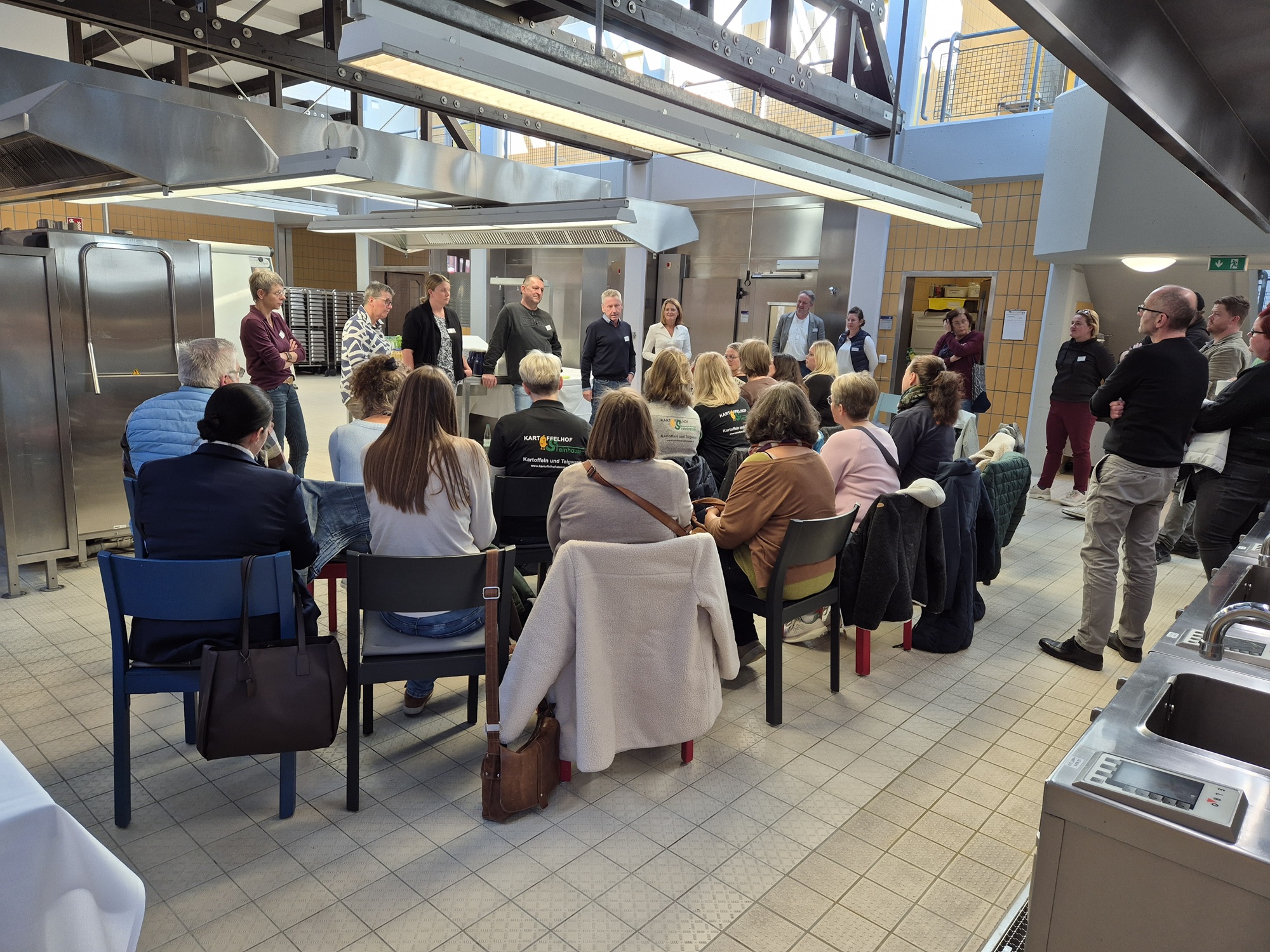Teilnehmende sitzen auf Stühlen in einer Großküche. Man sieht sie von hinten. Vor ihnen steht das Küchenteam der St. Elisabeth-Stiftung und erzählt.