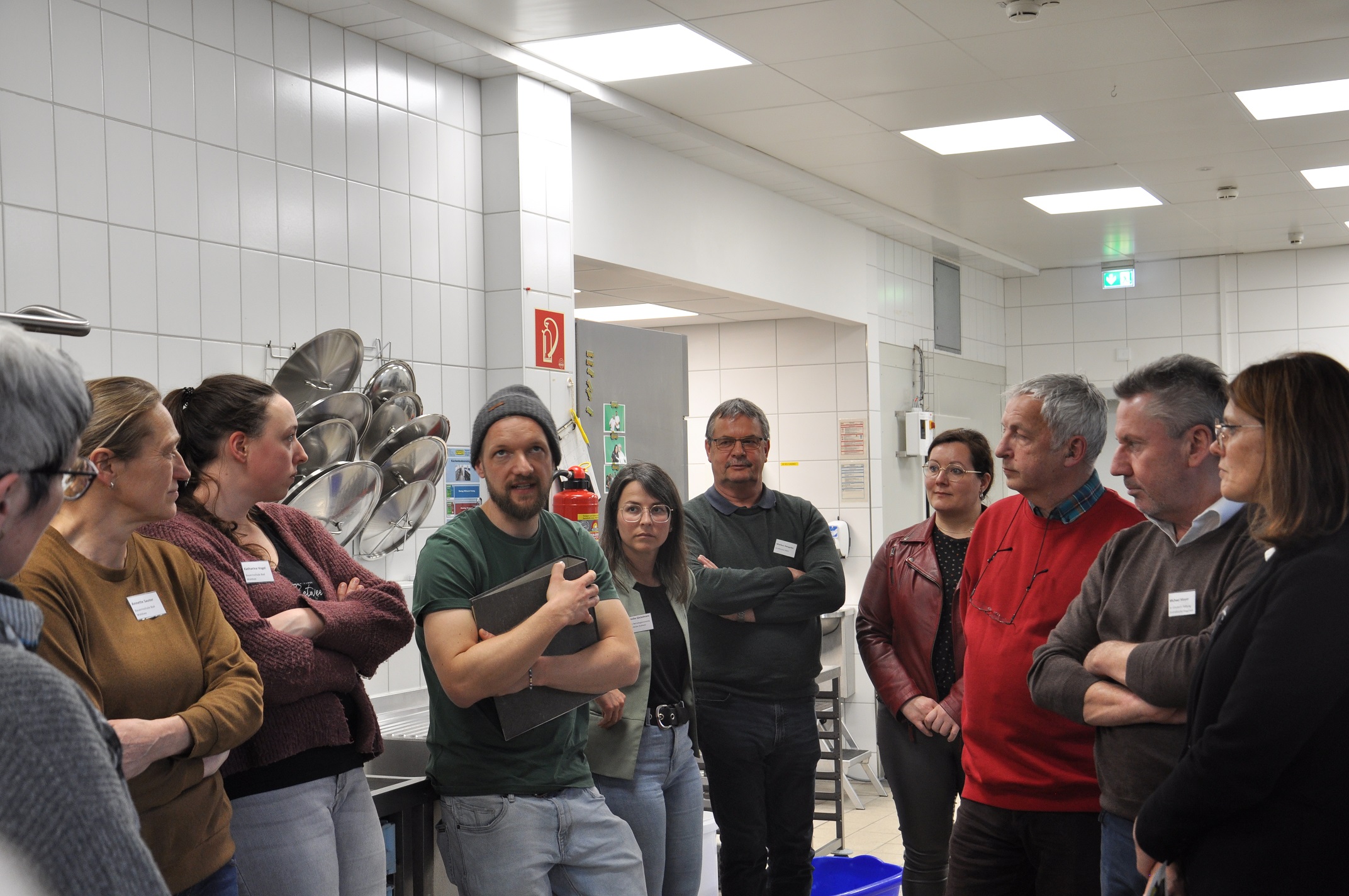 David Appenmaier steht in der Mitte der um ihn platzierten Teilnehmenden in der Großküche und berichtet aus der Praxis